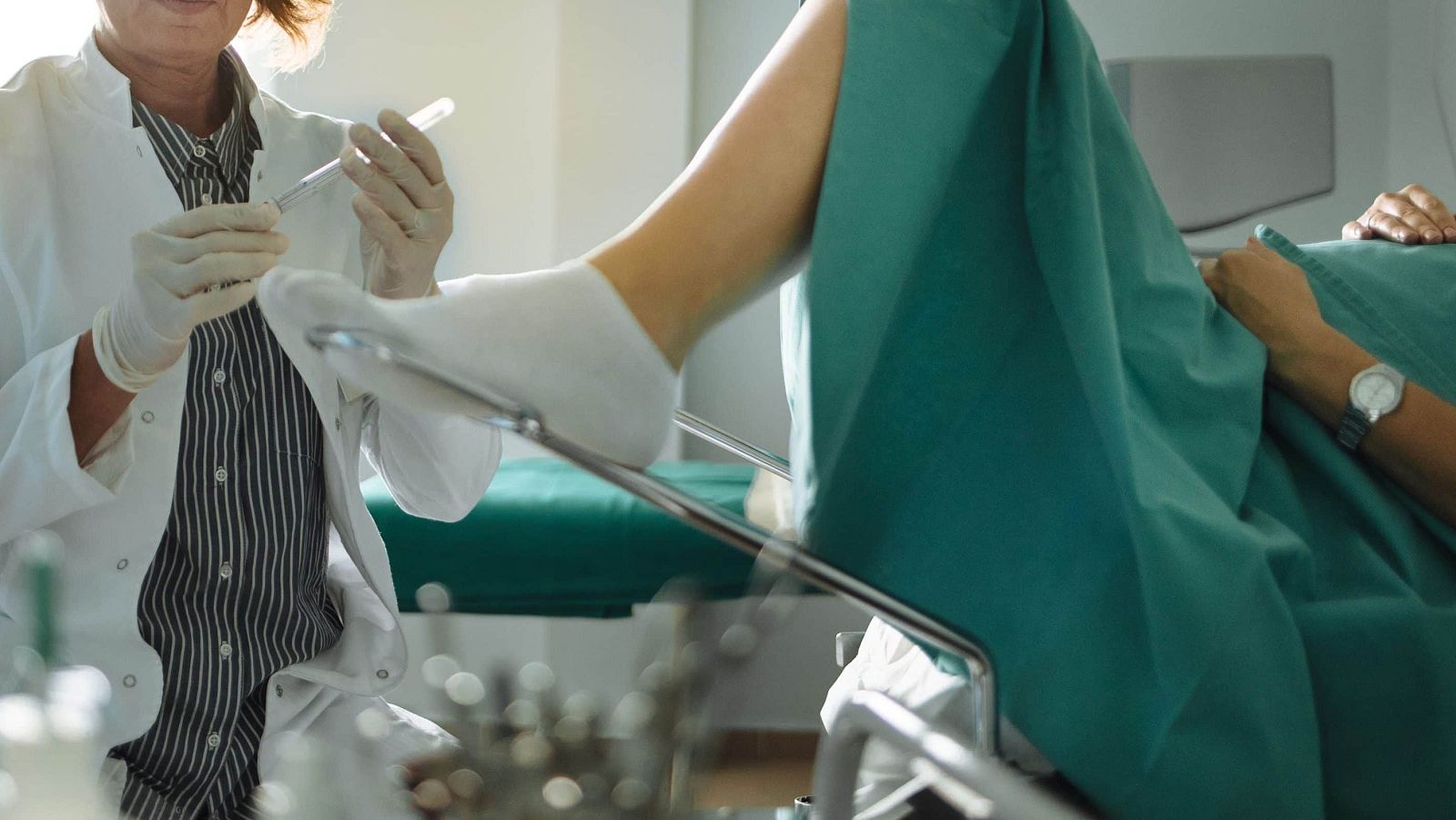 Doctora con bata blanca y guantes sostiene un vial frente a una paciente recostada en una camilla de exploración ginecológica con una sábana verde sobre sus piernas.