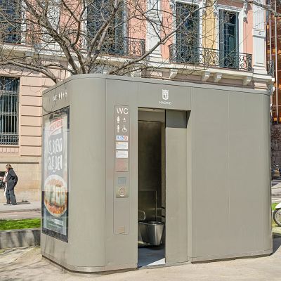 Cabina de aseo público gris en Madrid con el logo municipal, cartel de \"WC\" y un anuncio de Mercadona que dice \"LO BUENO SALE BIEN\", en una calle peatonal con peatones y una moto.