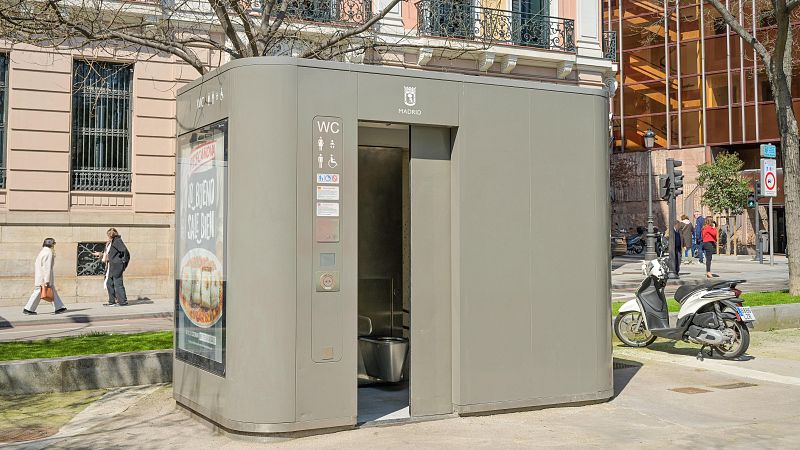 Cabina de aseo público gris en Madrid con el logo municipal, cartel de "WC" y un anuncio de Mercadona que dice "LO BUENO SALE BIEN", en una calle peatonal con peatones y una moto.