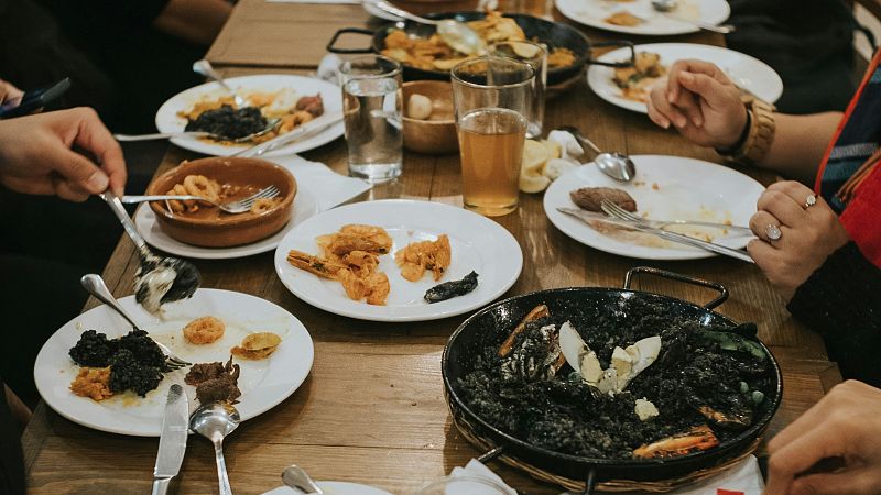 Varias personas comparten una comida espa�ola en una mesa de madera con paella de arroz negro, calamares fritos y tapas en cazuelas de barro.
