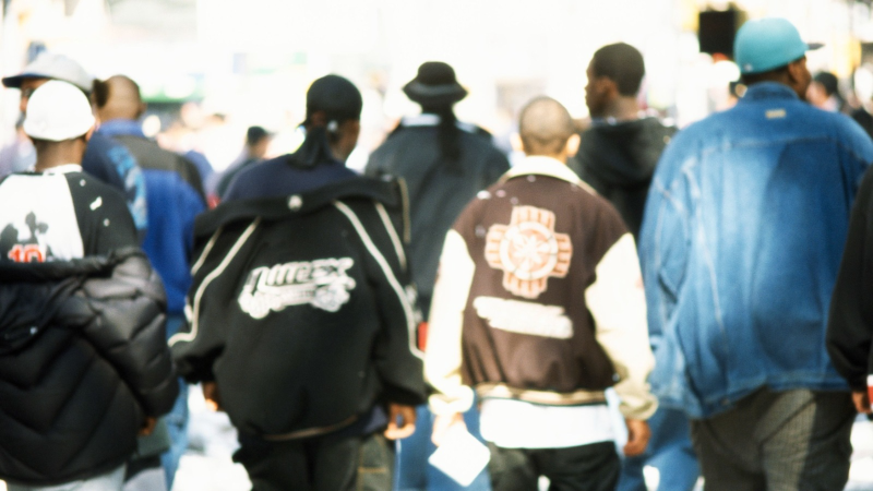 J�venes de espaldas con ropa urbana, incluyendo chaquetas acolchadas, universitarias y vaqueras con gorras, caminando por una calle soleada.