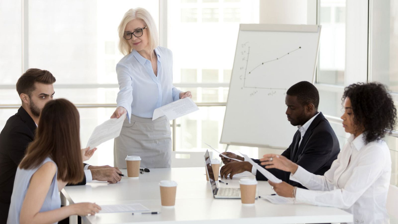 Una mujer rubia con gafas reparte folios a un grupo de profesionales multiculturales sentados en una mesa de oficina con port�tiles, caf�s y una pizarra de fondo.