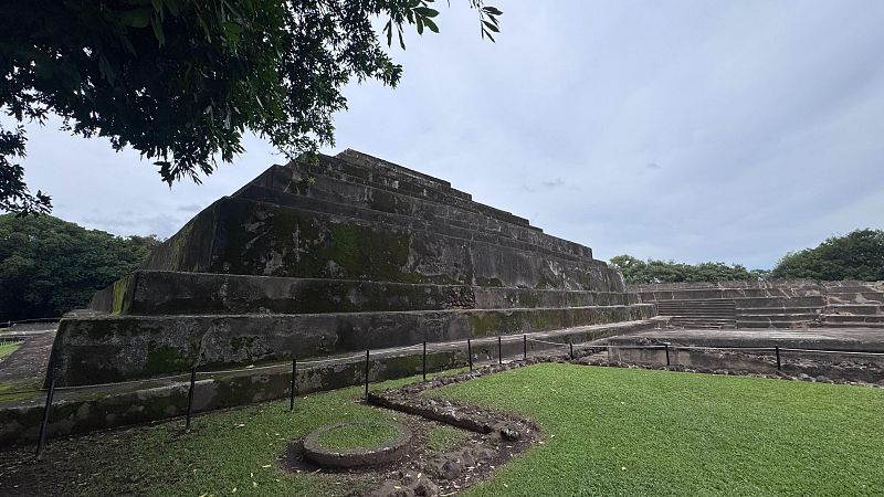 Pirámide escalonada de piedra gris del sitio arqueológico de Tazumal en El Salvador, rodeada de césped verde con una barrera protectora y árboles bajo un cielo nublado.