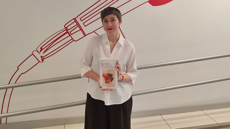 La escritora Elena Medel posa de pie sosteniendo su libro "Todos mis pecados y la mitad de mi dolor" frente a una pared blanca con un dibujo rojo de un micrófono.