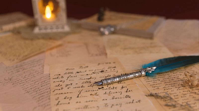 Pluma de escribir azul turquesa sobre cartas antiguas escritas a mano, iluminada por la c�lida luz de una vela dentro de un farolillo blanco al fondo.