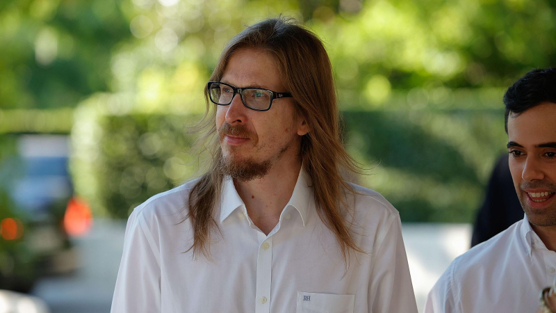 El pol�tico Pablo Fern�ndez Santos, con cabello largo, gafas y camisa blanca, posa en un exterior luminoso junto a otro hombre joven en un fondo desenfocado.