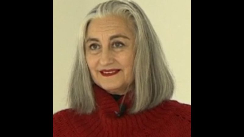 Retrato de la artista Pilar Albarracín con jersey rojo de cuello alto, micrófono de solapa y cabello gris plateado sobre un fondo claro.