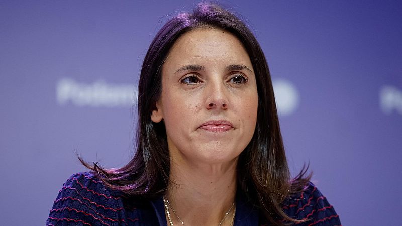 Retrato en primer plano de la política Irene Montero con expresión seria, vistiendo una prenda azul oscuro sobre un fondo morado desenfocado con la palabra "Podemos" sutilmente visible.