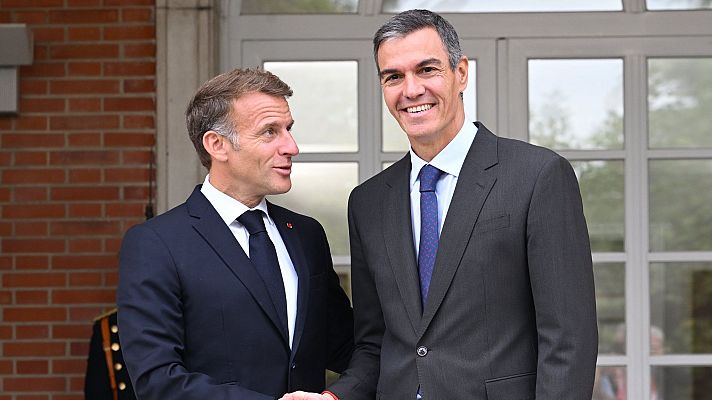 El presidente francés Emmanuel Macron y el presidente español Pedro Sánchez se saludan sonrientes con un apretón de manos durante un encuentro oficial frente a un edificio institucional.