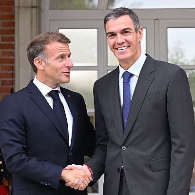 El presidente francés Emmanuel Macron y el presidente español Pedro Sánchez se saludan sonrientes con un apretón de manos durante un encuentro oficial frente a un edificio institucional.
