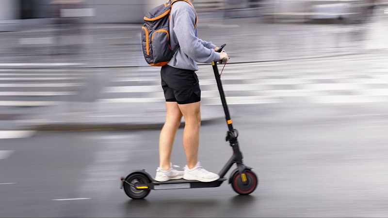 Persona en movimiento sobre un patinete el�ctrico negro, vistiendo sudadera gris, pantalones cortos y mochila gris con naranja, con un fondo urbano difuminado por la velocidad.