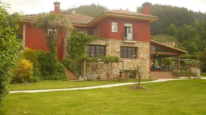 Fachada del hotel rural Alavera, con planta baja de piedra, piso superior rojo terracota y un amplio jardín delantero rodeado de frondosa vegetación.