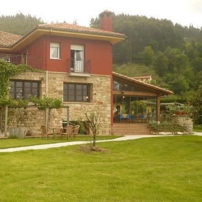 Fachada del hotel rural Alavera, con planta baja de piedra, piso superior rojo terracota y un amplio jardín delantero rodeado de frondosa vegetación.