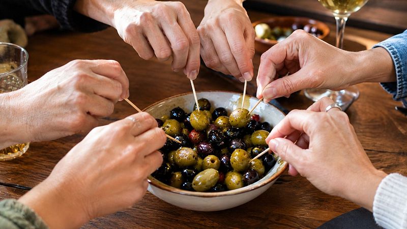 Varias personas comparten un cuenco de aceitunas ali�adas sobre una mesa de madera, pinch�ndolas con palillos en un ambiente c�lido y distendido.