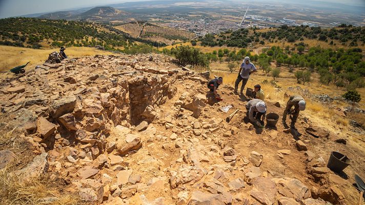 Arqueólogos trabajan en la excavación de una estructura circular de piedra en una ladera de Toledo, rodeados de herramientas y con un pueblo al fondo.