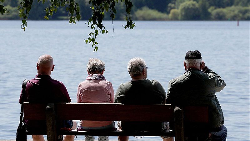 Cuatro personas mayores de espaldas, vestidas con ropa de abrigo y casual, contemplan sentadas en un banco de madera un tranquilo lago rodeado de naturaleza.