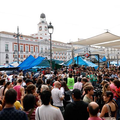 Multitudinaria protesta en la Puerta del Sol de Madrid, con un campamento improvisado y una persona en primer plano vistiendo un chaleco reflectante verde con el logotipo 'STOP'.