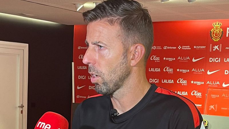 Jagoba Arrasate, con camiseta deportiva negra, habla en rueda de prensa frente a un micrófono de rne y un panel publicitario rojo con patrocinadores y el escudo del RCD Mallorca.