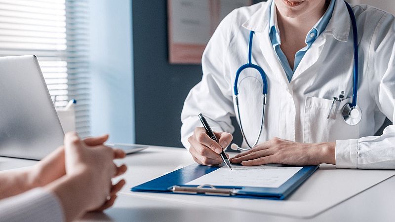 Un médico con bata blanca y estetoscopio escribe en una carpeta frente a un paciente cuyas manos están entrelazadas sobre la mesa de la consulta.