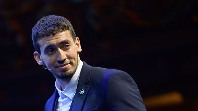 Mohamed El Amrani, con americana azul y micr�fono de diadema, sonr�e de perfil en un plano medio con fondo oscuro durante un evento.