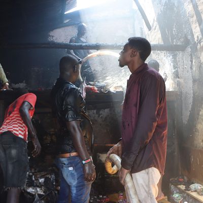 Hombres en Sudán trabajan entre escombros y humo dentro de un edificio calcinado, usando agua para apagar los restos del fuego bajo la luz que se filtra del techo.