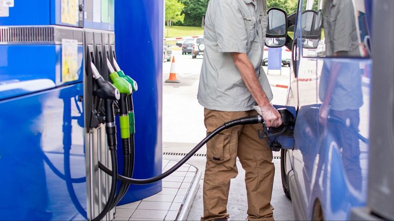 Un hombre con camisa gris y guante de plástico reposta combustible en una furgoneta azul junto a un surtidor en una estación de servicio exterior.