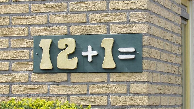 Placa verde sobre pared de ladrillo con la operaci�n matem�tica "12 + 1 =" en relieve de colores amarillo y blanco, utilizada de forma original para evitar el n�mero 13 en una vivienda.