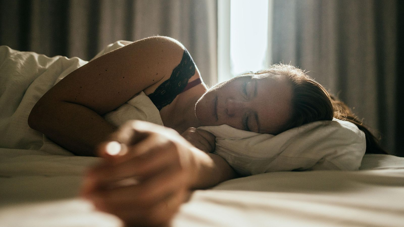 Mujer durmiendo de lado en cama, cabello casta�o, sujetador de encaje negro, tatuaje en brazo. Luz natural diurna.