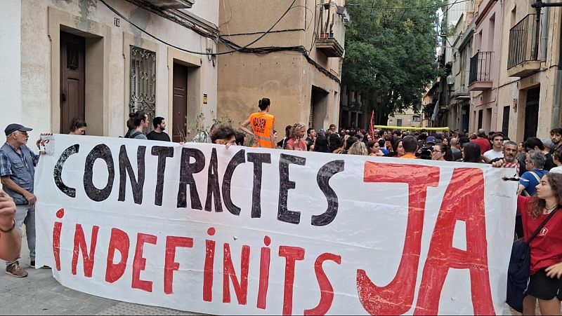 Manifestantes en una calle estrecha sostienen una gran pancarta que exige �CONTRACTES INDEFINITS JA�, junto a un miembro de un sindicato con chaleco reflectante.