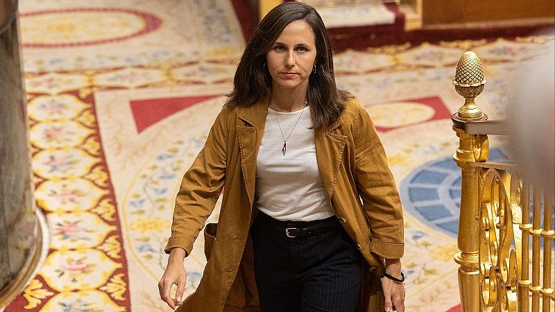 La pol�tica Ione Belarra camina con expresi�n seria por el Congreso de los Diputados, vistiendo una gabardina mostaza sobre una camiseta blanca y pantalones oscuros.