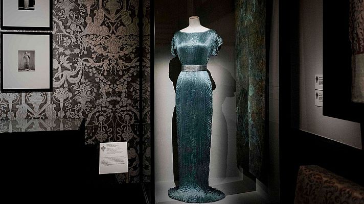 Vestido Delphos de seda plisada turquesa, diseñado por Mariano Fortuny, expuesto en una vitrina iluminada del Museo del Traje junto a fotografías históricas y paneles informativos.