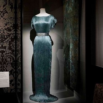 Vestido Delphos de seda plisada turquesa, diseñado por Mariano Fortuny, expuesto en una vitrina iluminada del Museo del Traje junto a fotografías históricas y paneles informativos.