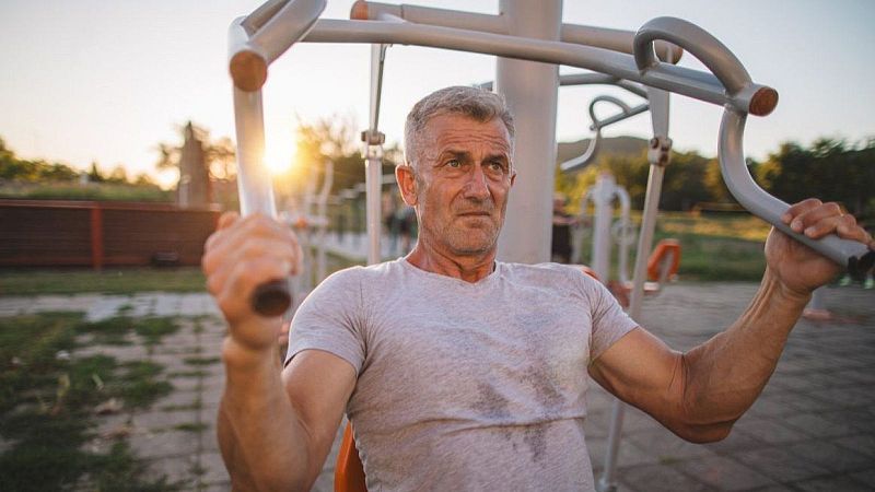 Hombre de mediana edad y cabello canoso se ejercita concentrado en una m�quina de gimnasio al aire libre en un parque bajo la c�lida luz del atardecer.