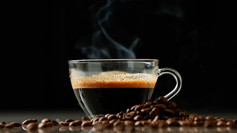 Taza de cristal transparente con caf� negro caliente y una densa capa de crema, rodeada de granos de caf� tostados sobre un fondo negro con vapor ascendente.
