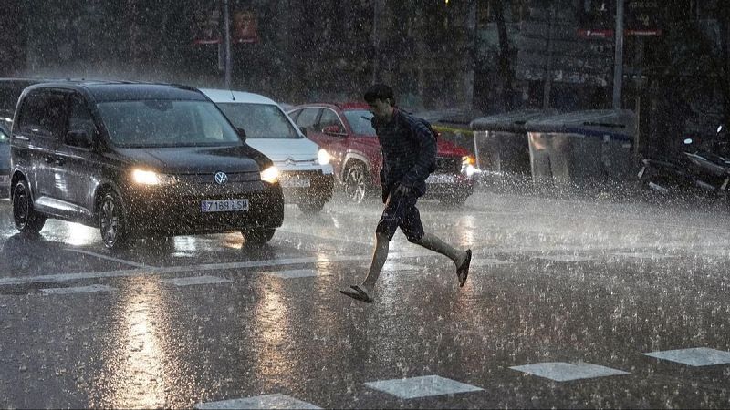 Un joven corre por un paso de peatones bajo una tormenta junto a una furgoneta Volkswagen con matrícula 7189 LSM y otros vehículos con los faros encendidos.