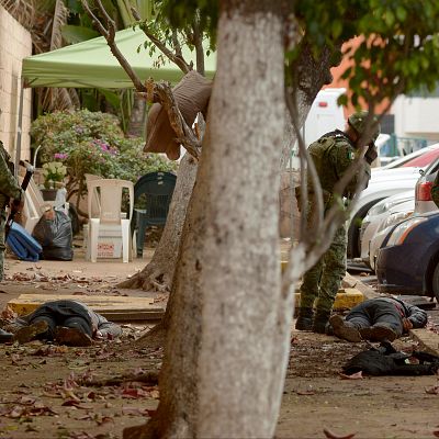 Soldados del Ejército Mexicano con uniformes de camuflaje y armas largas custodian una escena con dos personas tendidas en el suelo en una zona urbana.