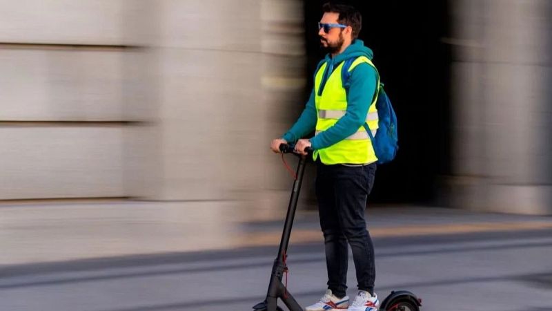 Hombre con barba, sudadera azul y chaleco reflectante amarillo conduce un patinete eléctrico negro a gran velocidad por una calle de la Comunidad Valenciana.