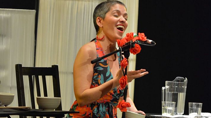Una mujer con vestido estampado canta expresivamente ante un micrófono adornado con rosas rojas en un escenario teatral con atrezo.