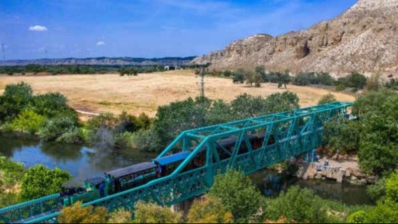Un hist�rico tren de vapor con vagones de madera cruza un puente met�lico verde sobre un r�o, rodeado de vegetaci�n y un paisaje �rido bajo un cielo azul.