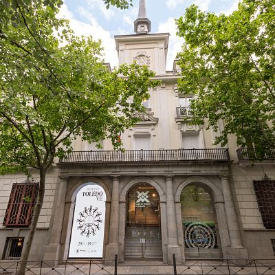 Fachada simétrica de la Casa de México en Madrid, destacando tres arcos de piedra con un cartel de la exposición \"TOLEDO VE\" de Francisco Toledo y frondosos árboles a los lados.