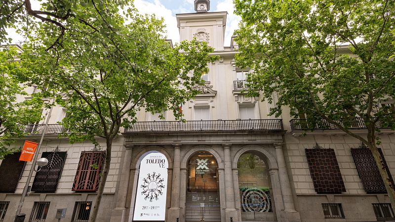 Fachada simétrica de la Casa de México en Madrid, destacando tres arcos de piedra con un cartel de la exposición "TOLEDO VE" de Francisco Toledo y frondosos árboles a los lados.