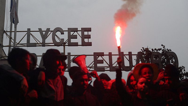 Jóvenes protestan frente al Lycée Paul Éluard en Francia, destacando uno que sostiene una bengala roja encendida y otro con un megáfono bajo un cielo nublado.
