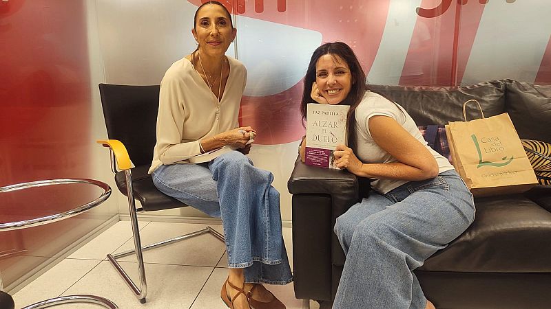 Paz Padilla y Marta González de Vega posan sonrientes en un estudio de RNE; Marta sostiene el libro "Madre!" de Paz junto a una bolsa de Casa del Libro.