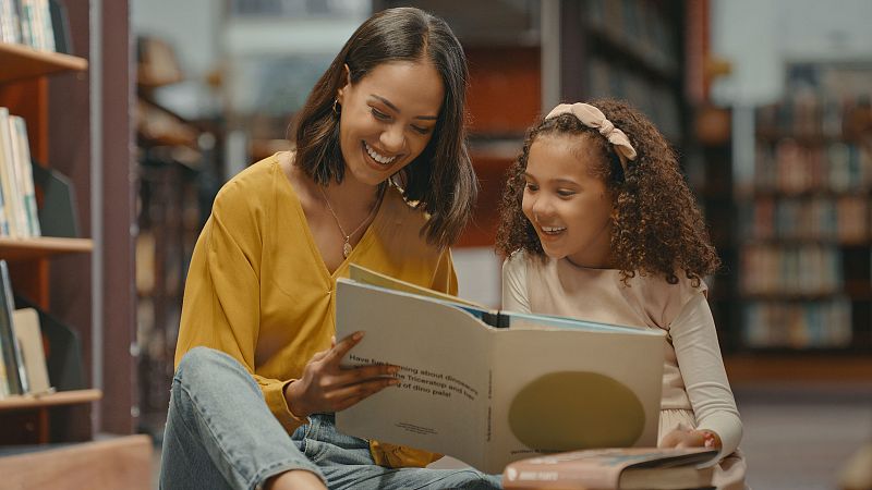 Una mujer con blusa amarilla y una niña con camiseta rosa leen un libro infantil sentadas en el suelo de una biblioteca, rodeadas de estanterías de madera.