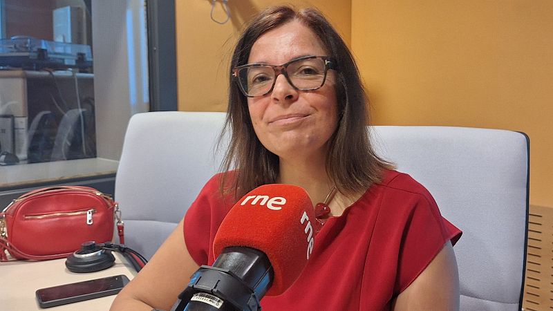 Margarida Romero sonríe en un estudio de Radio Nacional de España, sentada frente a un micrófono con el logo de "rne" y vistiendo una blusa roja.