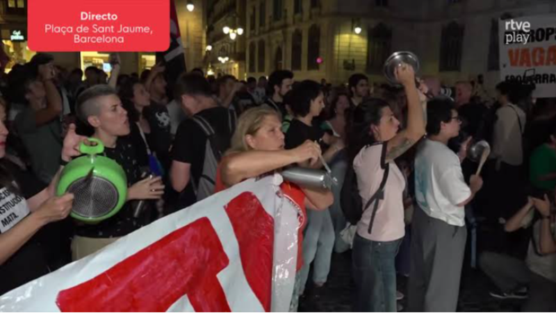 Protesta nocturna con cacerolada en una plaza iluminada de Barcelona, con personas golpeando utensilios de cocina y sosteniendo pancartas.