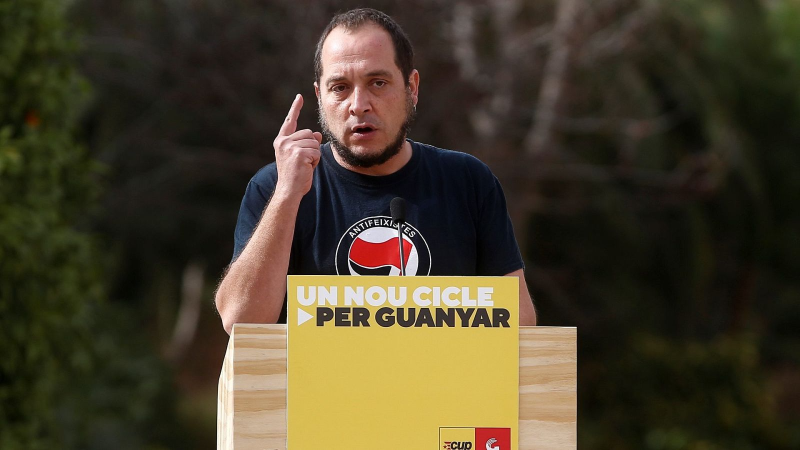 David Fern�ndez interviene tras un atril con un cartel amarillo que muestra el lema �UN NOU CICLE�, haciendo un gesto enf�tico con la mano.