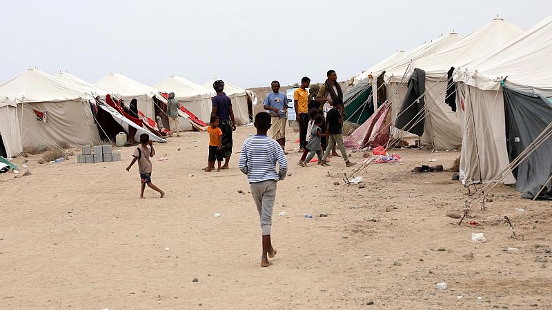 Campamento de refugiados en terreno desértico con tiendas blancas y personas caminando por el pasillo central de tierra.