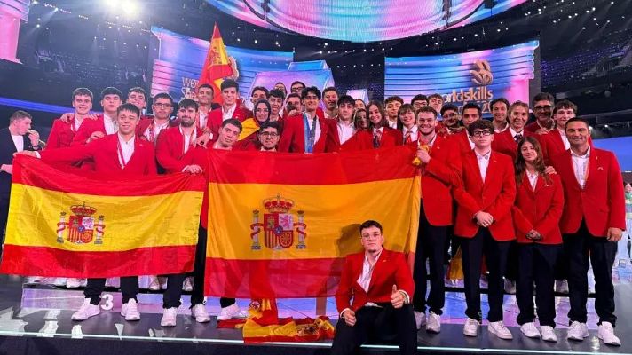 Delegación española posa con americana roja y banderas de España en el escenario iluminado de WorldSkills.