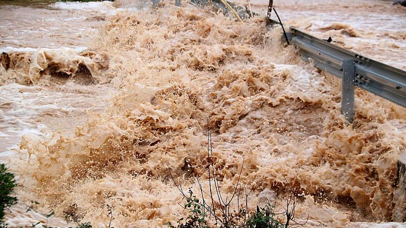 Una riada de agua turbia y marr�n desborda con fuerza una carretera, golpeando una barrera de seguridad met�lica situada a la derecha.